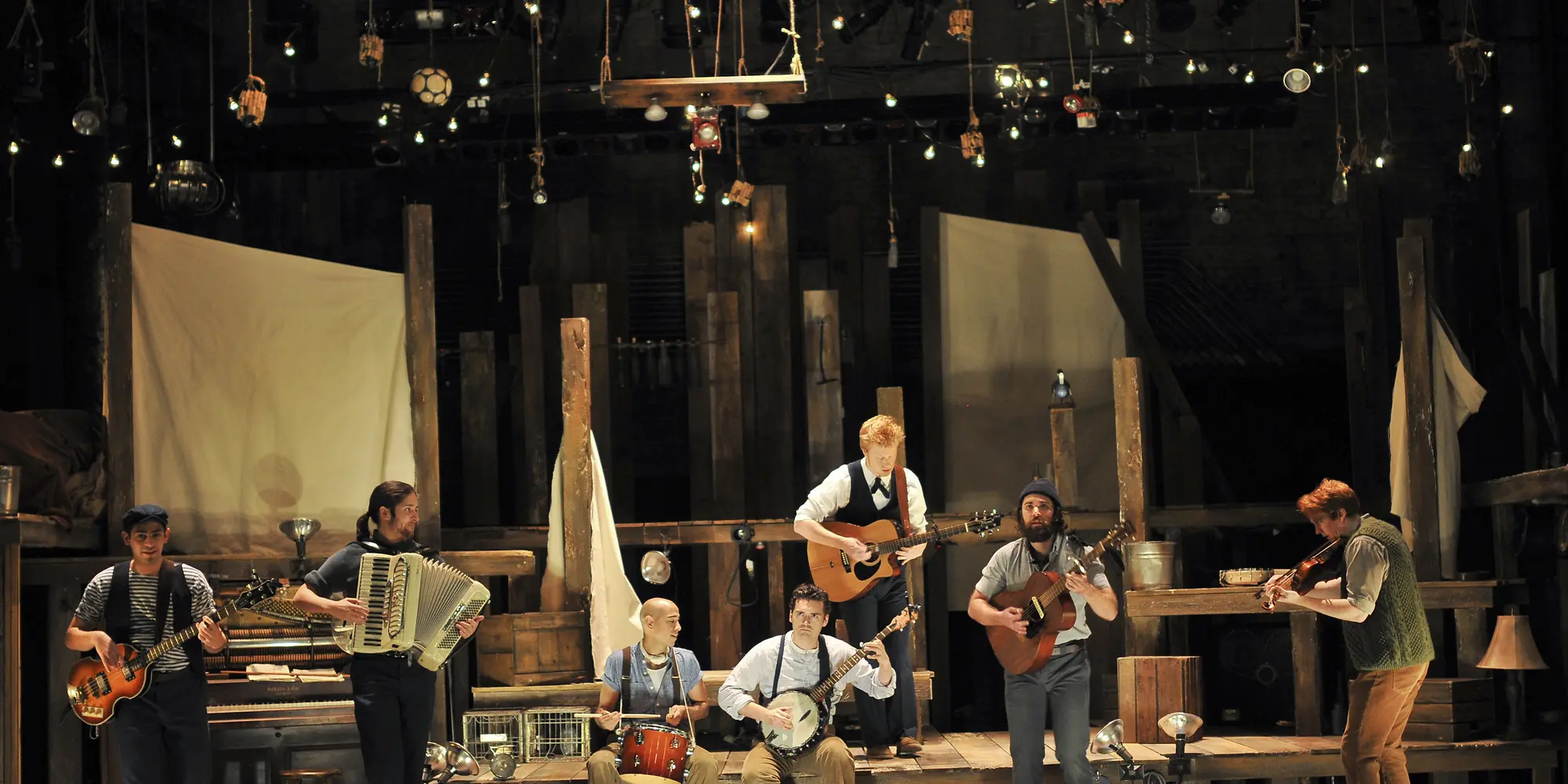 Seven men dressed as sailors on a steps of a wooden deck set on a theater stage.