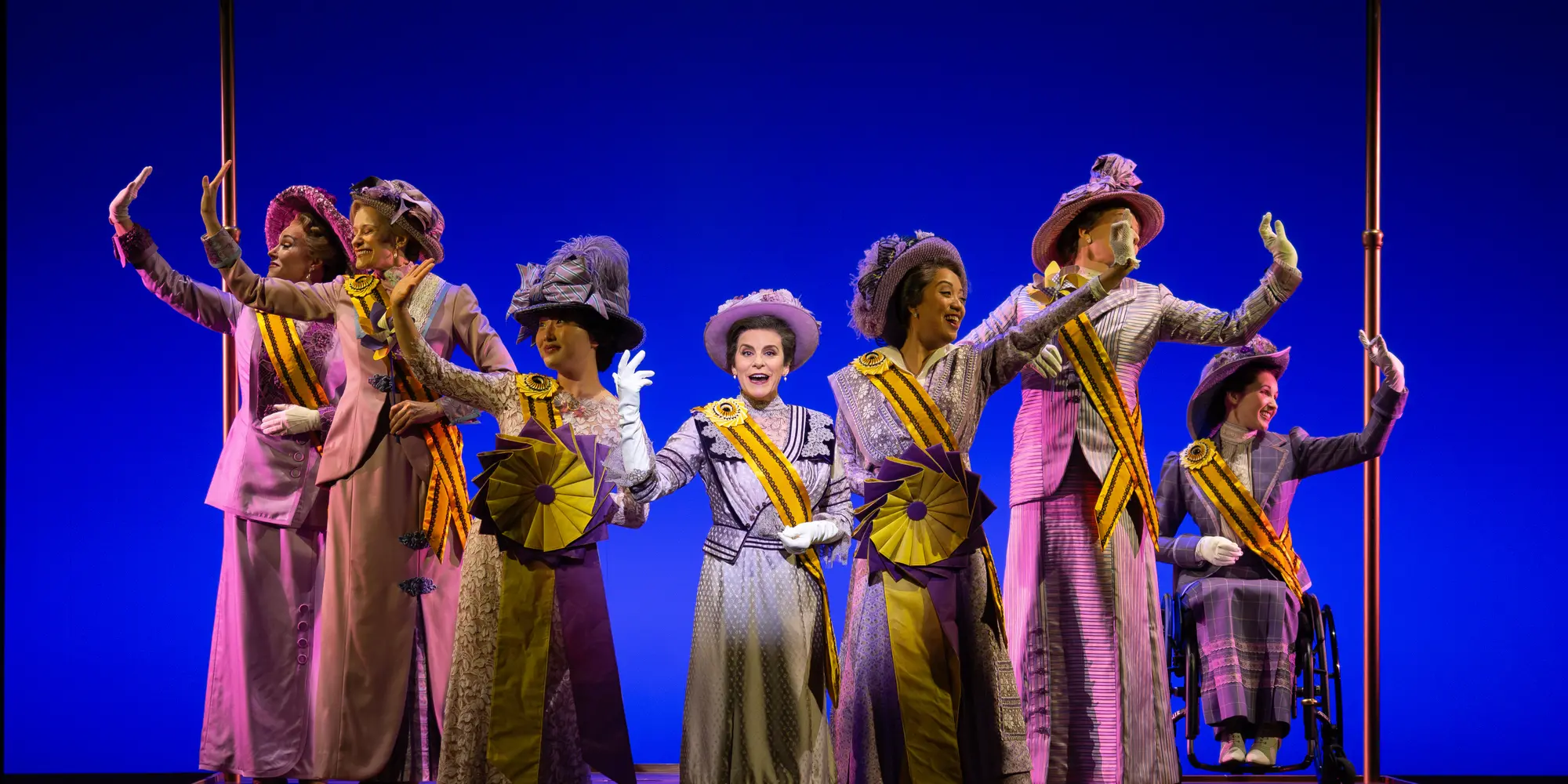 On a stage, six women in 1920s-era clothes and suffrage sashes stand on a smaller stage with a yellow banner above them that says, "National American Women Suffrage Association 1913," as each of them wave to the side, except for the woman in the middle who sings to the audience with a spotlight on her.