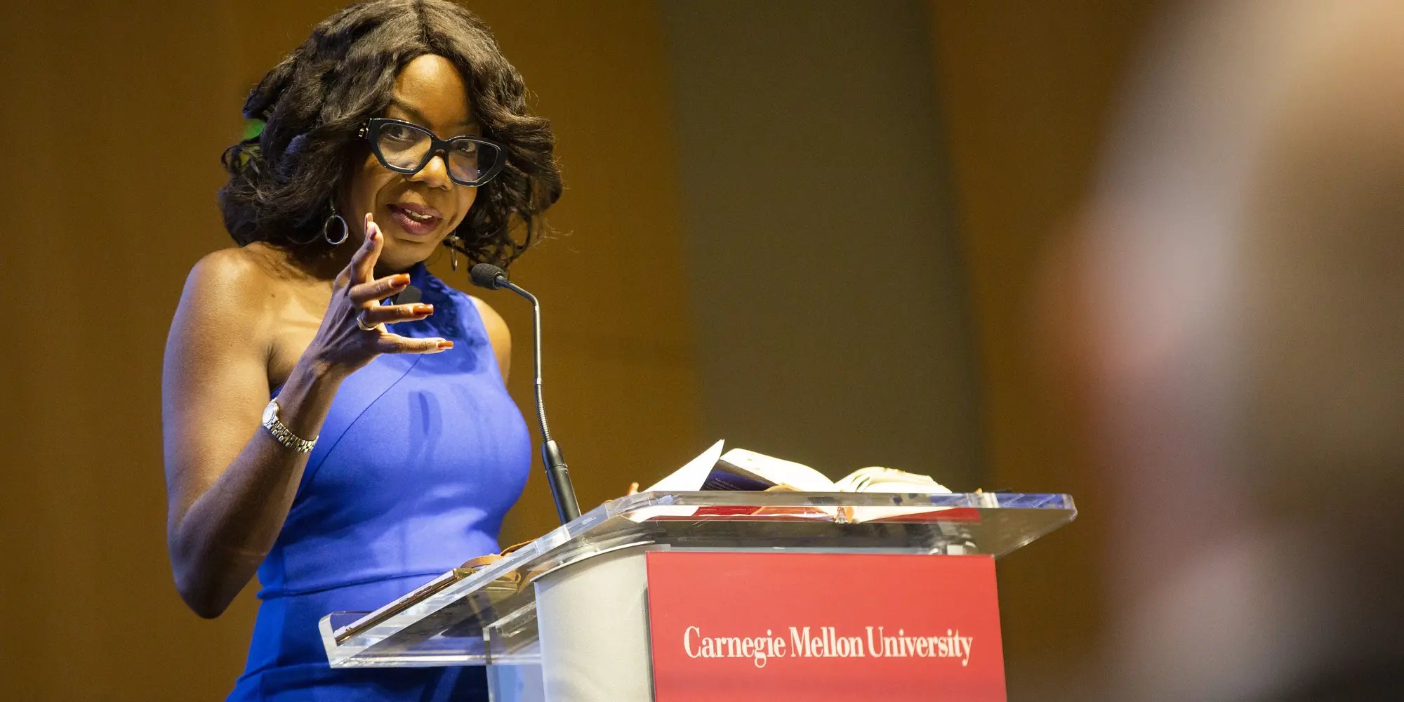 A Black woman with black-framed glasses, curled hair and hoop earrings wearing a blue one-shoulder dress, gestures toward the audience while standing at a lecturn with a red Carnegie Mellon University sign attached to the front. 