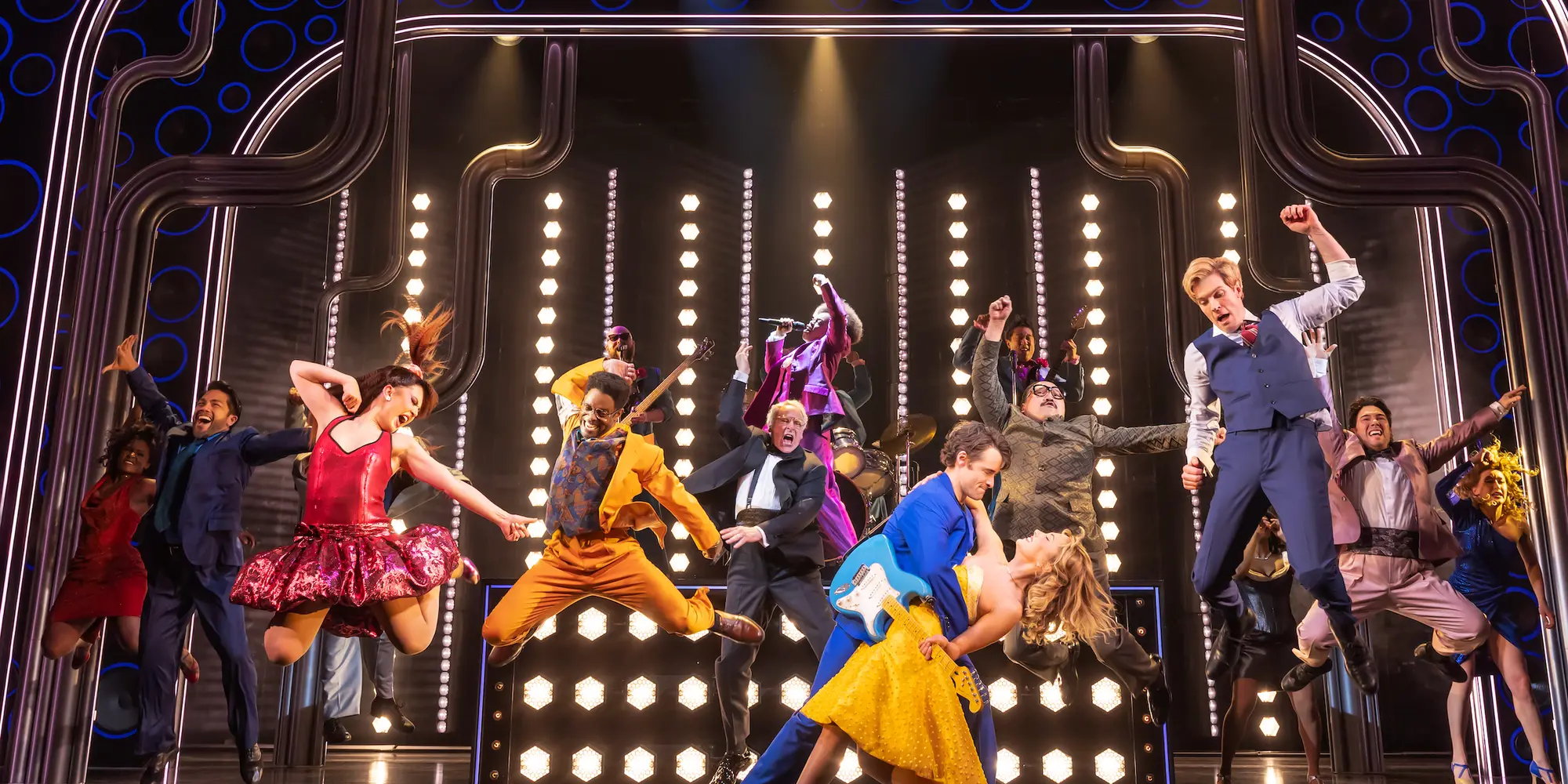 On a stage, a man in a blue suit with a guitar on his back dips a blonde woman in a yellow dress, meanwhile behind them, a cast of about a dozen people in brightly colored '80s clothing are frozen mid-jump in a dance number in front of a set comprised of rows of white lights. 