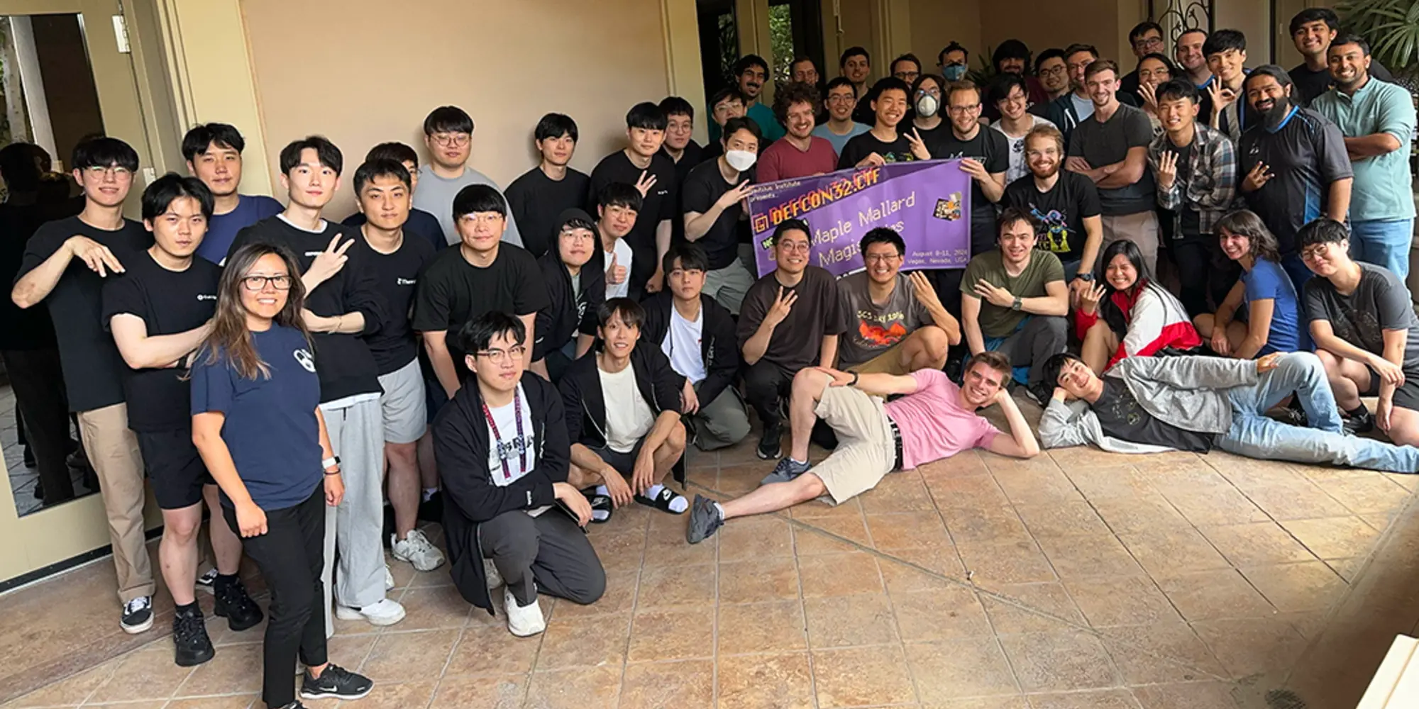 A large group of about 50 people, most wearing black T-shirts and holding up three fingers to signify the team's third win, pose for a photo with a banner including the team name.