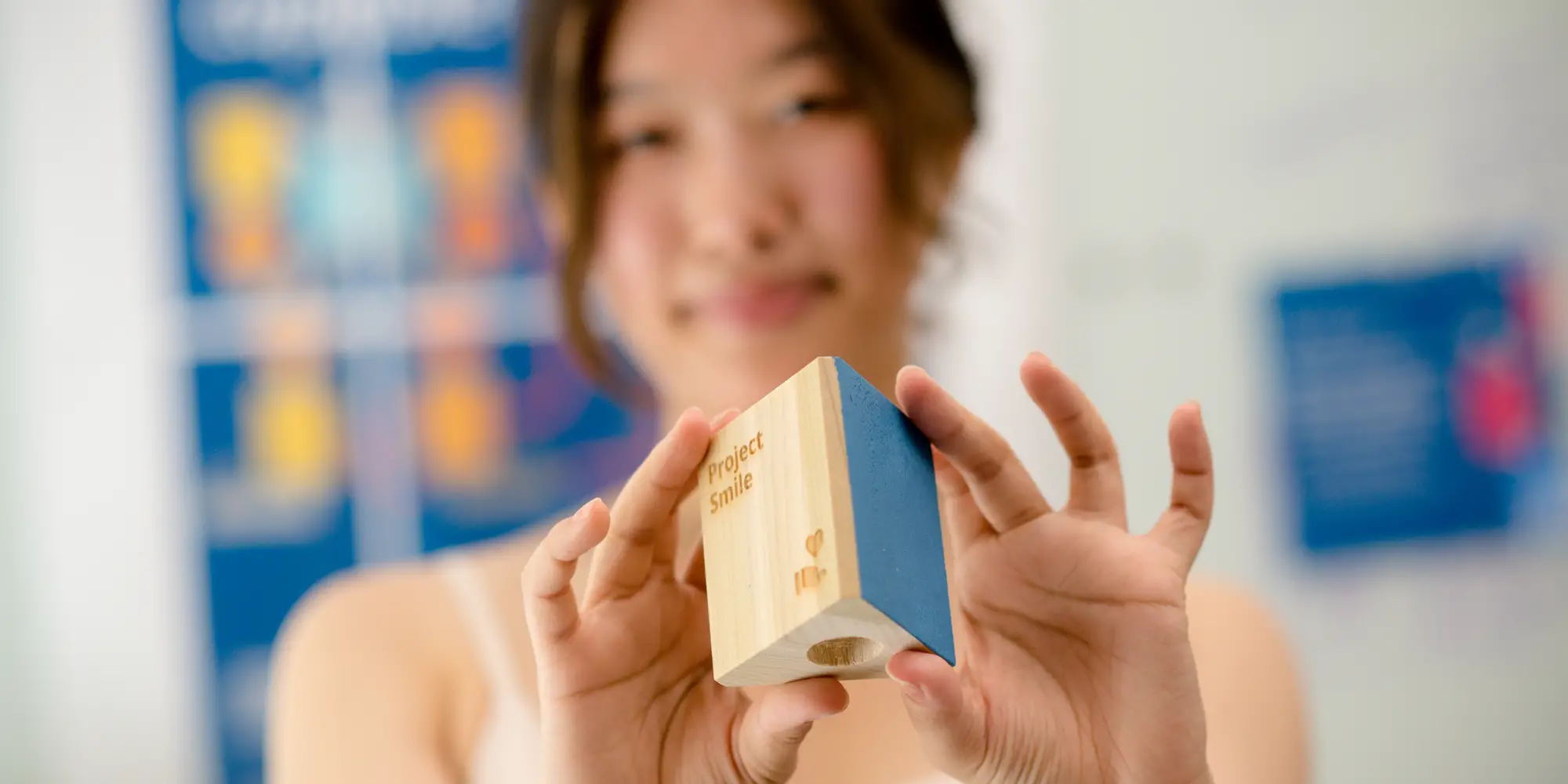 A woman out-of-focus in the background holds up a wooden block in-focus in the foreground.