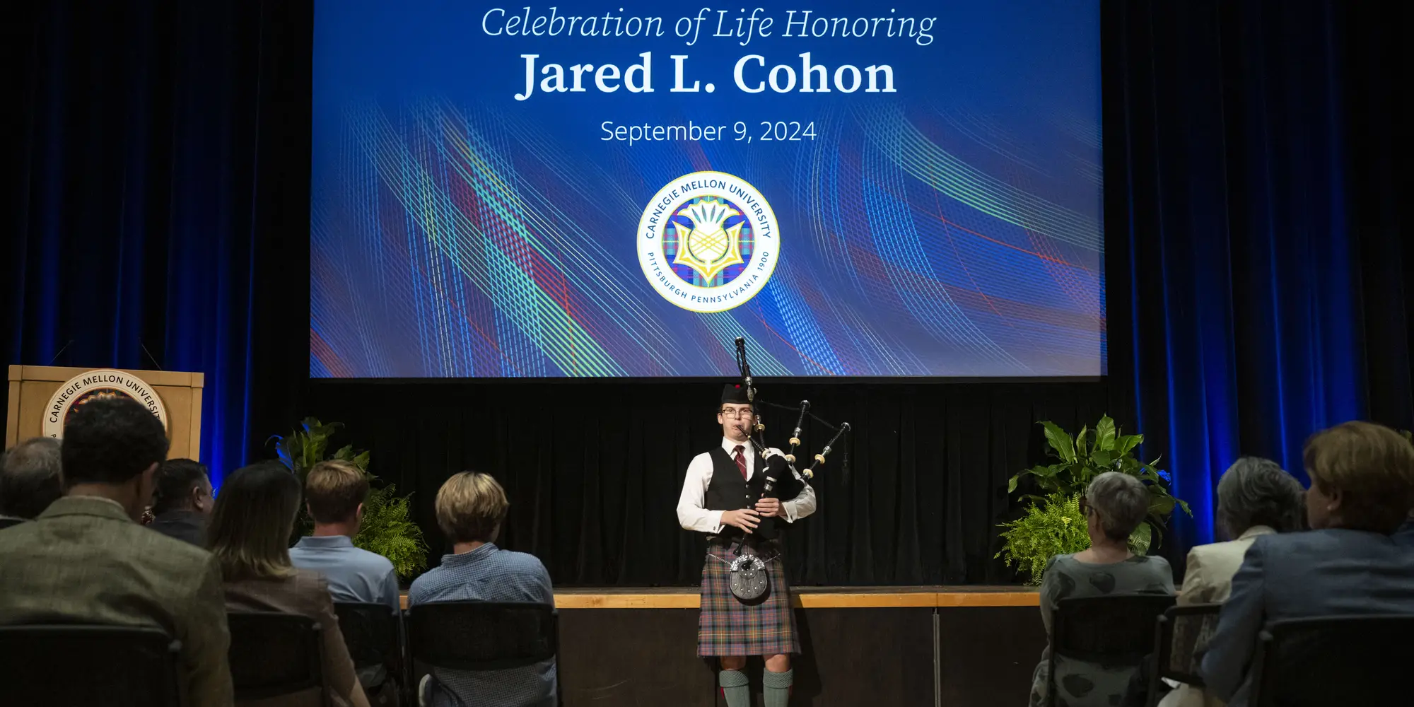 A man stands in front of a stage playing bagpipes dressed in traditional Scottish garb, including a kilt. Behind him, a screen displays a slide that says, "Celebration of Life Honoring Jared L. Cohan - September 9, 2024"