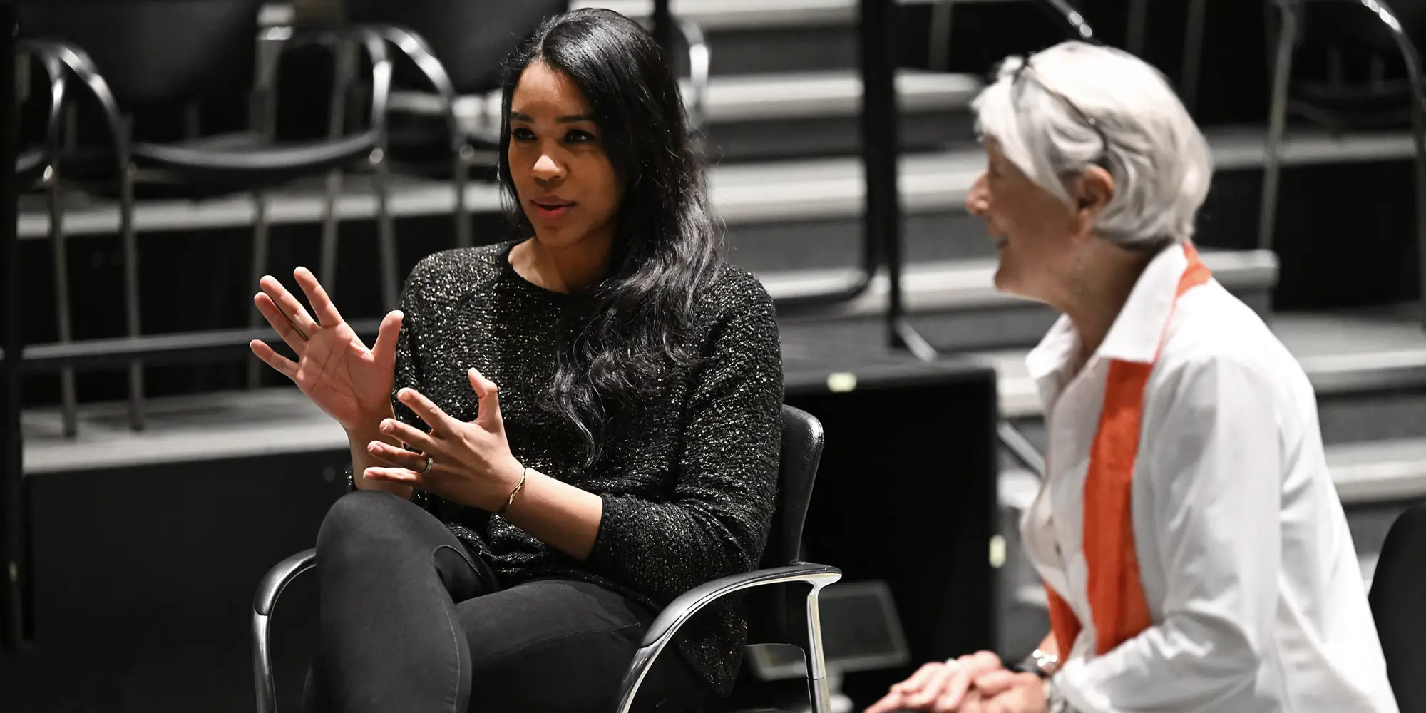 Two women seated in chairs look toward the left and speak to a crowd that is out of frame. The woman on the left is gesturing with her hands.