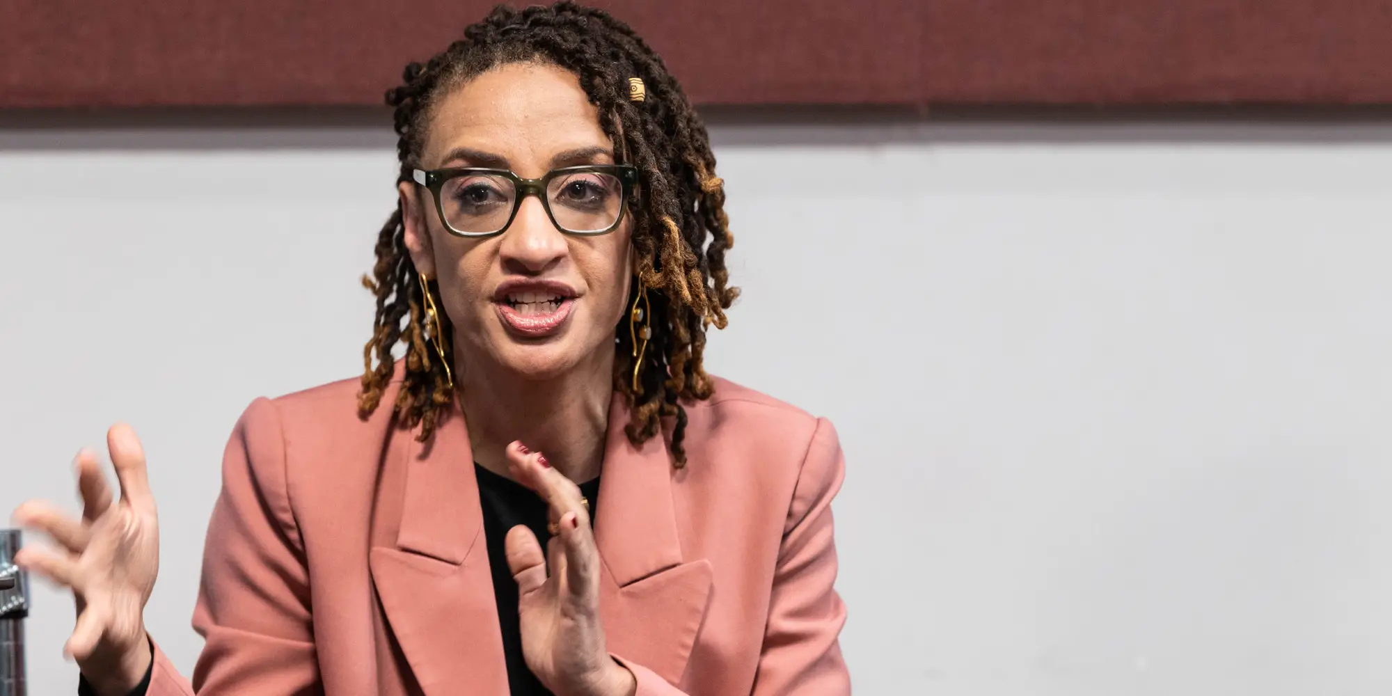 A woman with glasses wearing a salmon-colored coat gestures and talks