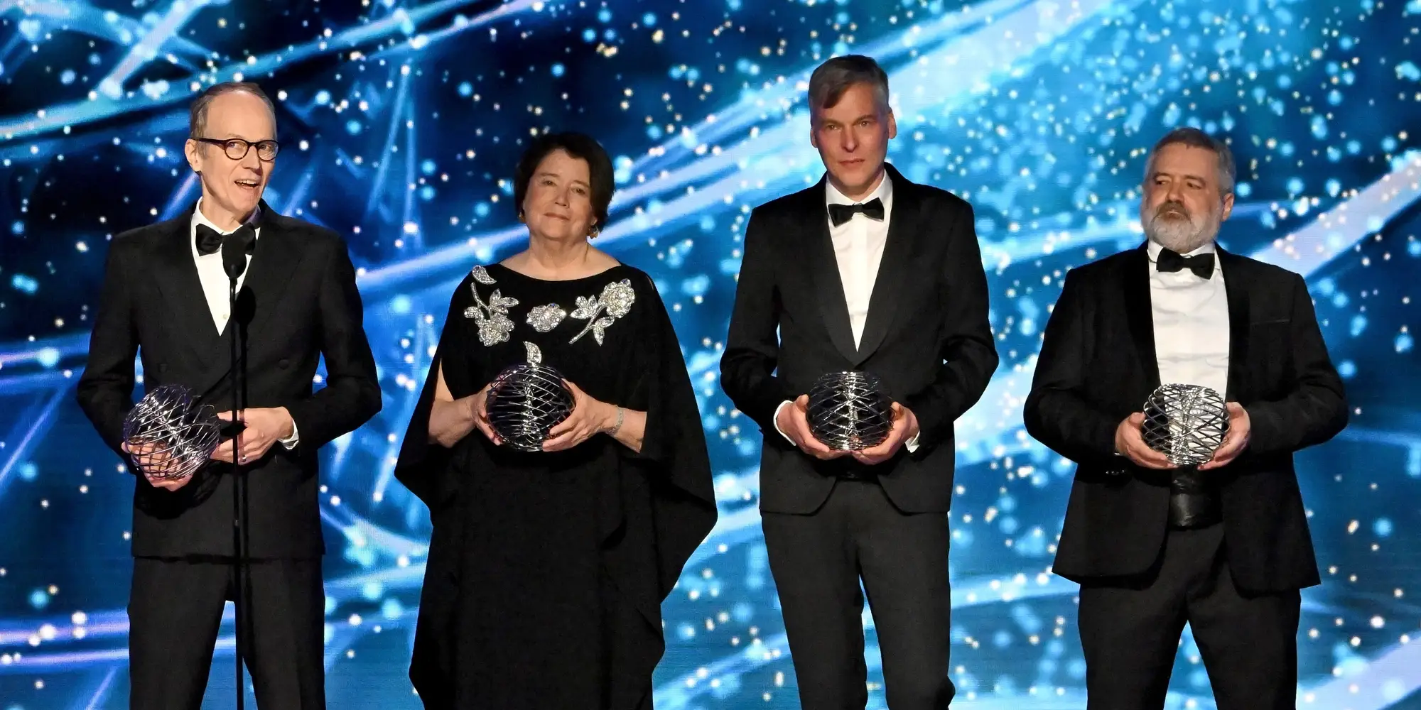 Three men and one woman hold spherical wire sculptures on a stage. The background is blue and filled with flashes of light.
