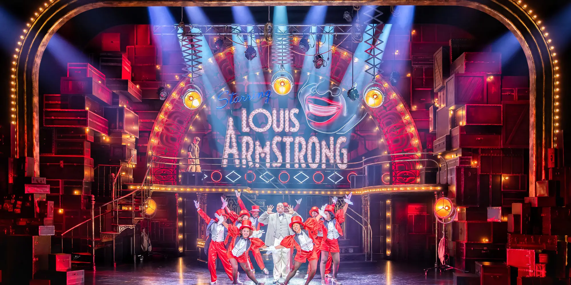 Stage and proscenium of a theater with sign on back wall that says "Louis Armstrong" and beams of light shining down on to cast members in red and silver sequined formal outfits in a small group.