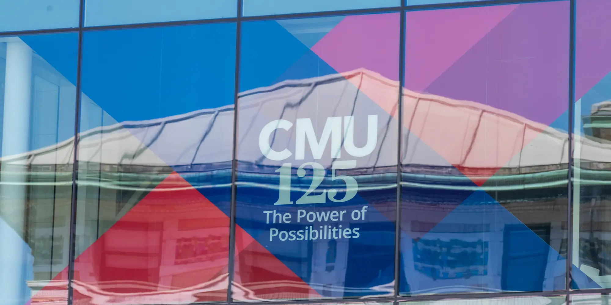 A picture of a window reflecting a building. In the window is a red and blue banner for CMU's 125th celebration. 