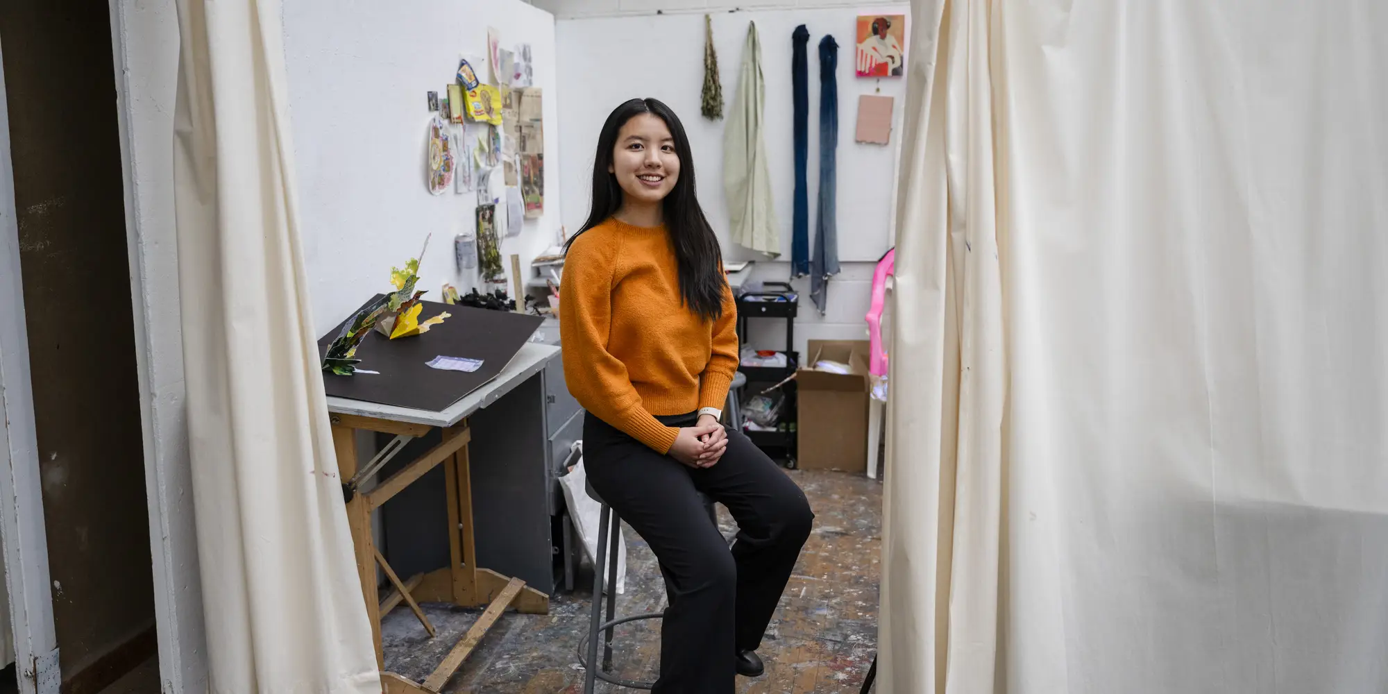 A woman with long black hair sits on a stool in front of an art studio with colorful art on the walls and a floor-to-ceiling curtain next to her.