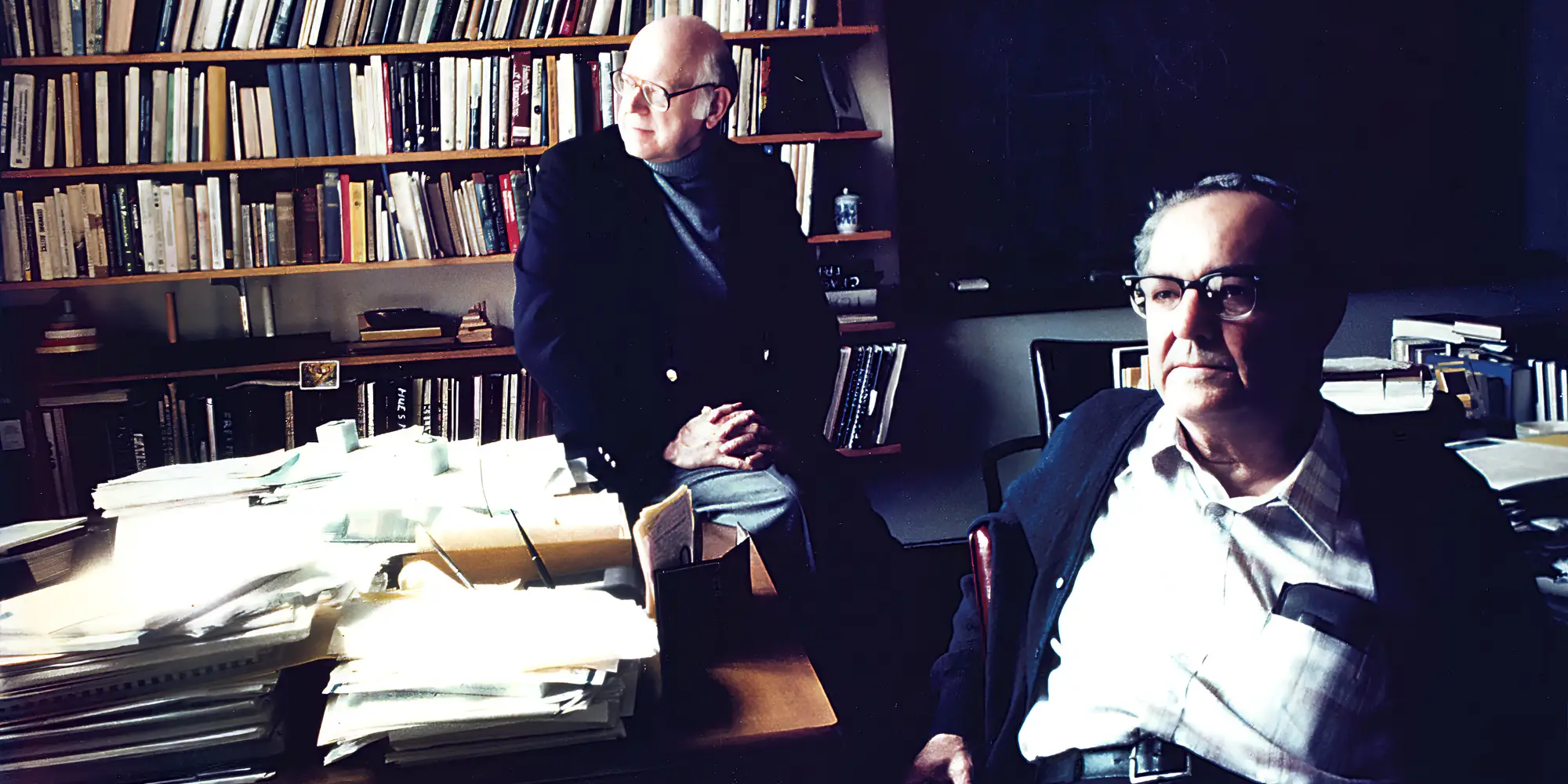 Herb Simon and Allan Newell sit in an office surrounded by books and papers. 
