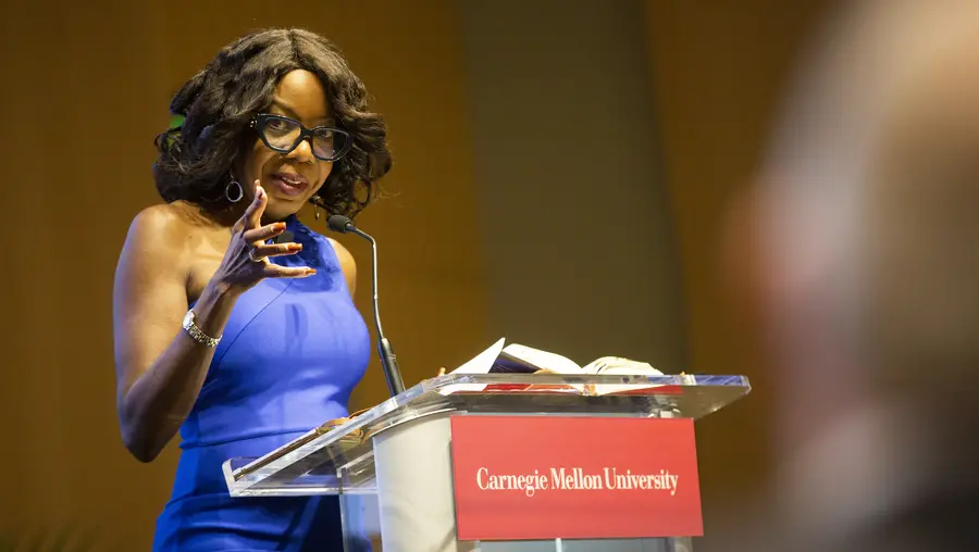 A Black woman with black-framed glasses, curled hair and hoop earrings wearing a blue one-shoulder dress, gestures toward the audience while standing at a lecturn with a red Carnegie Mellon University sign attached to the front. 