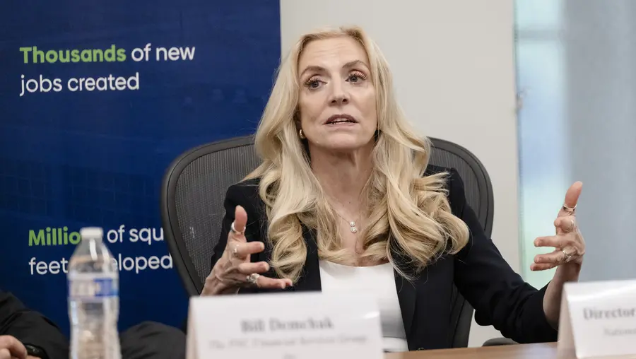 A white woman with long wavy blonde hair sits at a table behind a name placard with her hands raised to gesture while speaking.