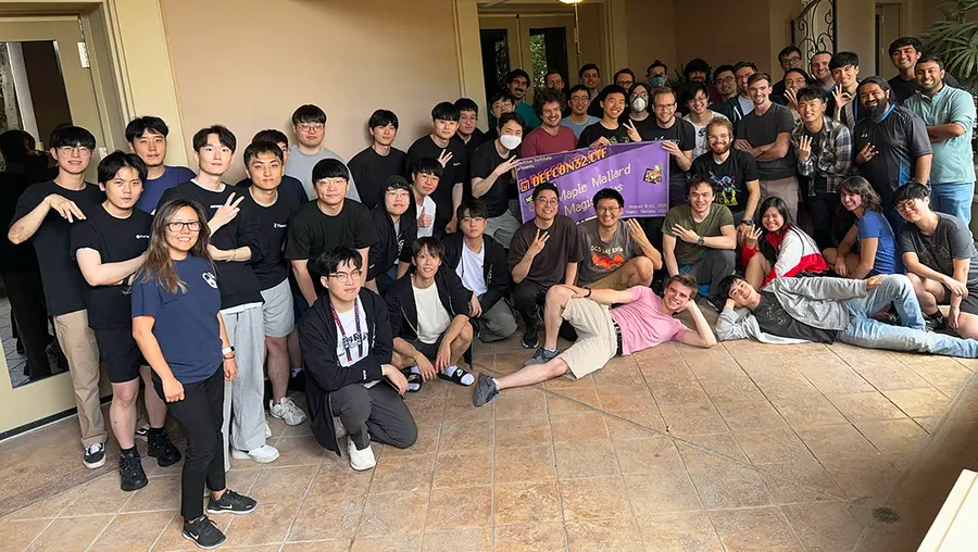 A large group of about 50 people, most wearing black T-shirts and holding up three fingers to signify the team's third win, pose for a photo with a banner including the team name.