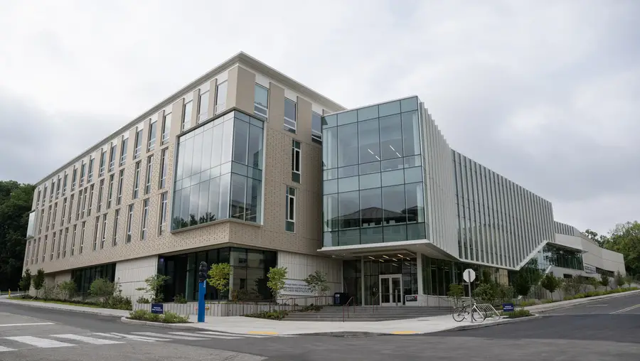 The exterior of a four-story building from a streetview of the intersection of roads where it sits, with a pattern of beige-colored brick covering the left side while a protruding cube of glass windows encompasses most of the right side.