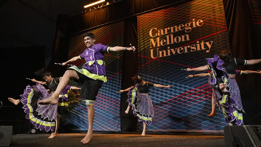 Five people in purple and green costumes kick their foot together as part of a dance group on a stage with a backdrop behind them that says 