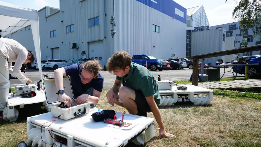 Two students assemble a robot in front of a building labeled NREC