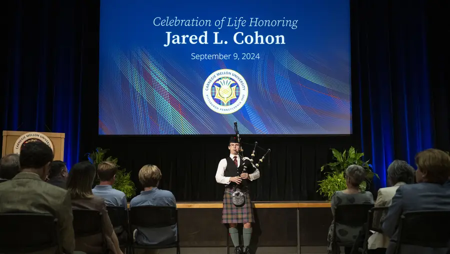 A man stands in front of a stage playing bagpipes dressed in traditional Scottish garb, including a kilt. Behind him, a screen displays a slide that says, "Celebration of Life Honoring Jared L. Cohan - September 9, 2024"