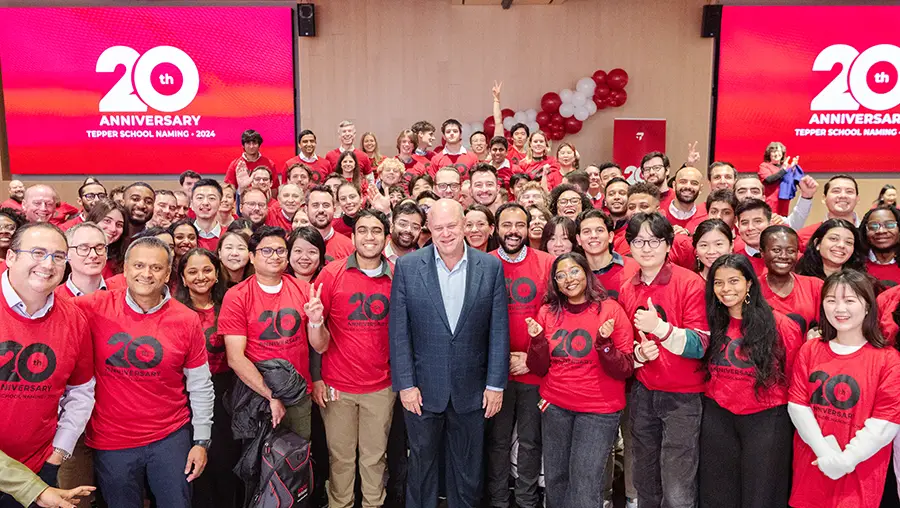 Large group of people all wearing the same red T-shirt, surrounded by a man in the business suit at the center. Large screens behind them are displying the same color red with white tex that says, "20th Anniversary - Tepper School naming 2024"