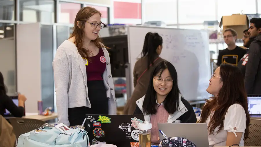 Woman standing at edge of table talks to two female students sitting at the table in front of their laptops.
