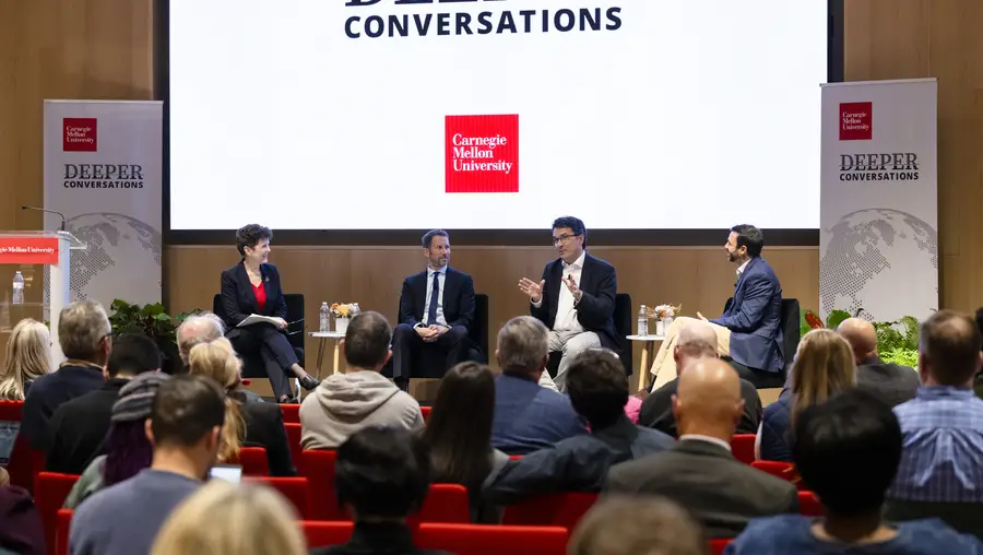 A group of four people on a stage sitting in chairs, in front of a screen that says "Deeper Conversations"