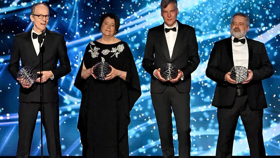 Three men and one woman hold spherical wire sculptures on a stage. The background is blue and filled with flashes of light.