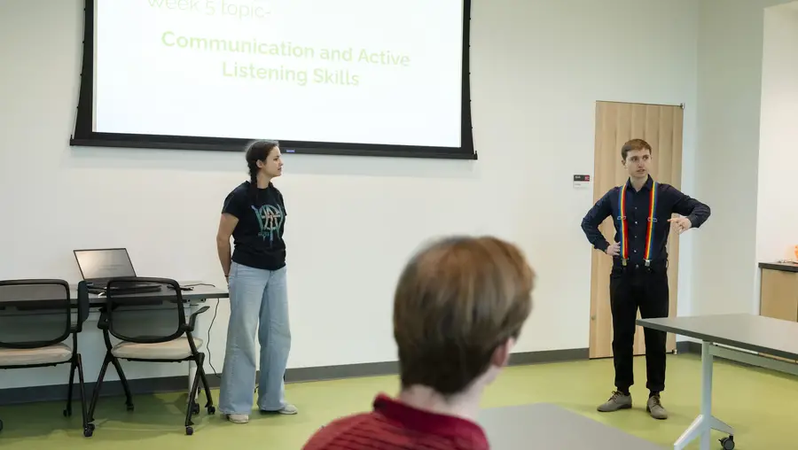 Two people stand at the front of a classroom underneath a projector screen talking while a student, listening and looking toward them, is framed in the foreground.