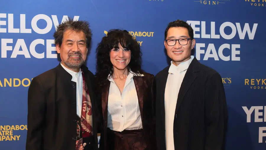 A woman stands between two men in front of a backdrop that reads "Yellow Face" on the left and right