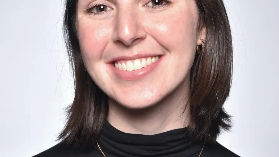 A head-and-shoulders portrait of a smiling woman with shoulder length brown hair and bangs in a black turtleneck wearing a small gold chain necklace.