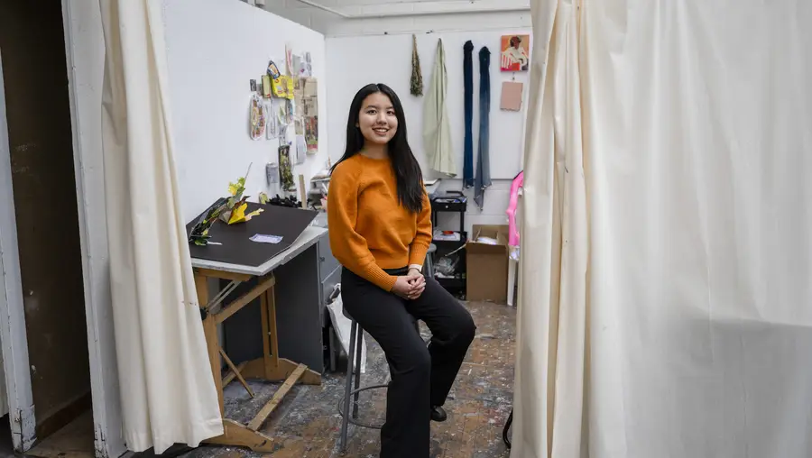 A woman with long black hair sits on a stool in front of an art studio with colorful art on the walls and a floor-to-ceiling curtain next to her.