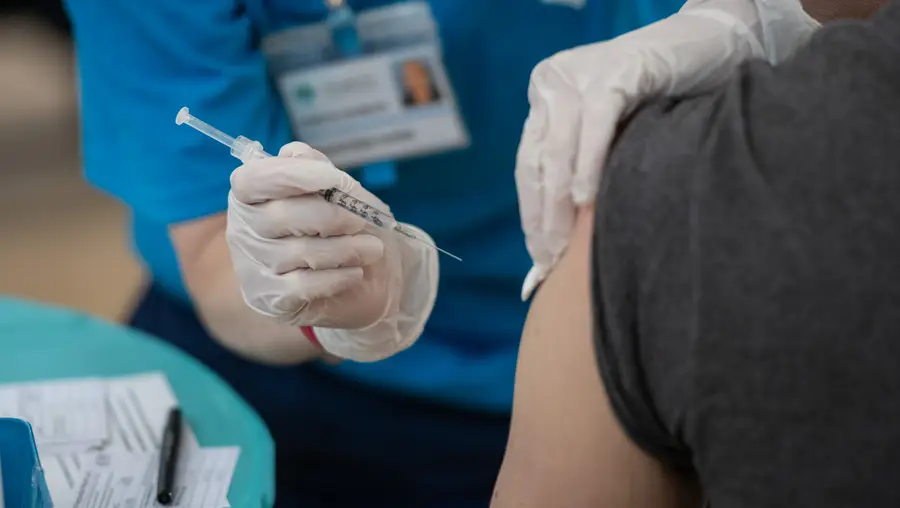 A close-up showing two gloved hands of a nurse holding a syringe of COVID-19 vaccine about to pierce the skin of someone's arm with their T-shirt sleeve rolled up.