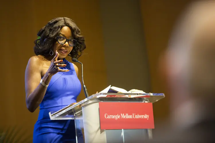 A Black woman with black-framed glasses, curled hair and hoop earrings wearing a blue one-shoulder dress, gestures toward the audience while standing at a lecturn with a red Carnegie Mellon University sign attached to the front. 