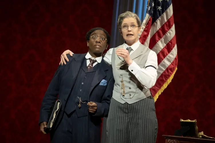 In 1920s-era costumes, a white woman dressed as a man with a moustache speaks with her arm around a Black woman dressed as a man, who looks reluctant to the companion's message. The pair stand in front of an American flag on a stand.