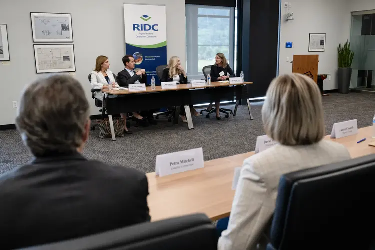 Three white women and one white man dressed in business attire talk at a table in the background, while the camera looks between the shoulders of a man and a woman sitting behind name placards at a table in the foreground.