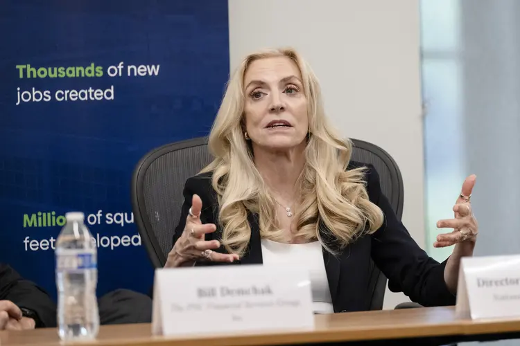 A white woman with long wavy blonde hair sits at a table behind a name placard with her hands raised to gesture while speaking.