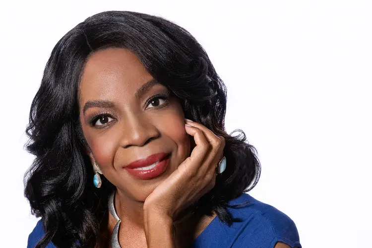 A Black woman with shoulder-length black hair in a blue dress sits in a wooden chair with her arm bent on the chair's arm and her chin resting in her hand.