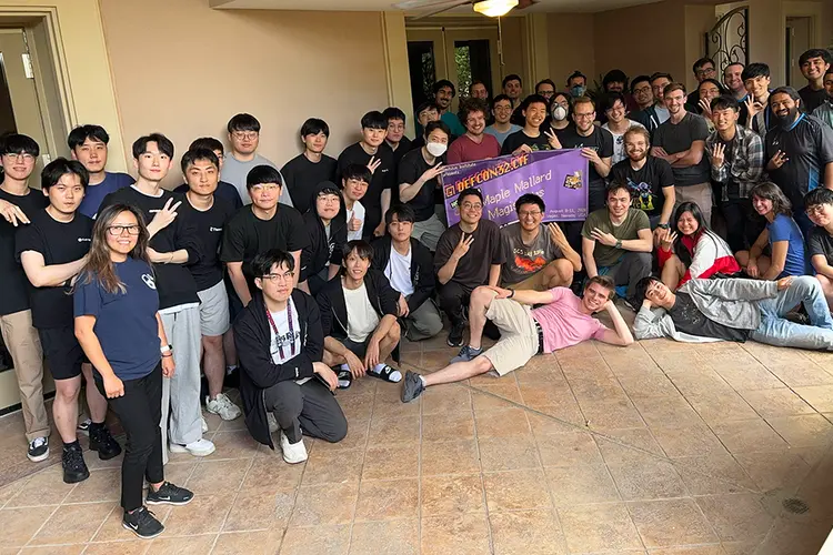 A large group of about 50 people, most wearing black T-shirts and holding up three fingers to signify the team's third win, pose for a photo with a banner including the team name.