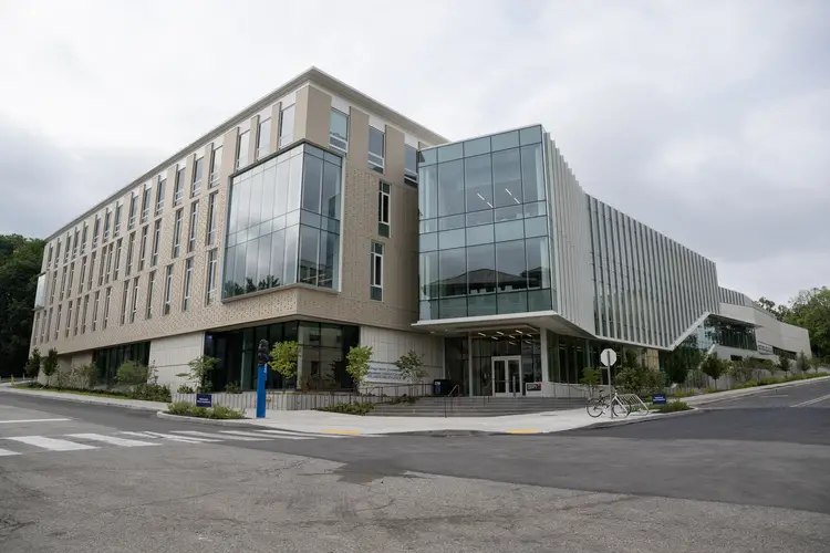 The exterior of a four-story building from a streetview of the intersection of roads where it sits, with a pattern of beige-colored brick covering the left side while a protruding cube of glass windows encompasses most of the right side.