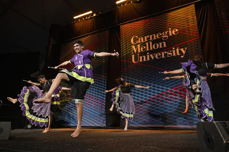 Five people in purple and green costumes kick their foot together as part of a dance group on a stage with a backdrop behind them that says 