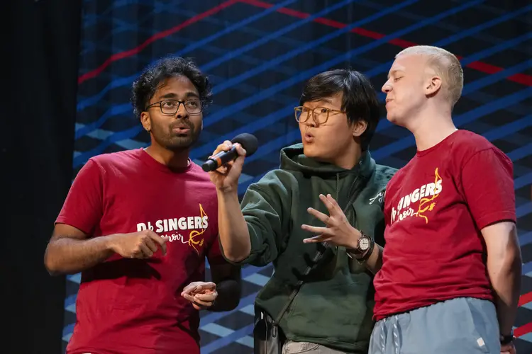 Three men make "o" shapes with their mouths as they sing. The man in the middle holds the microphone as they perform.