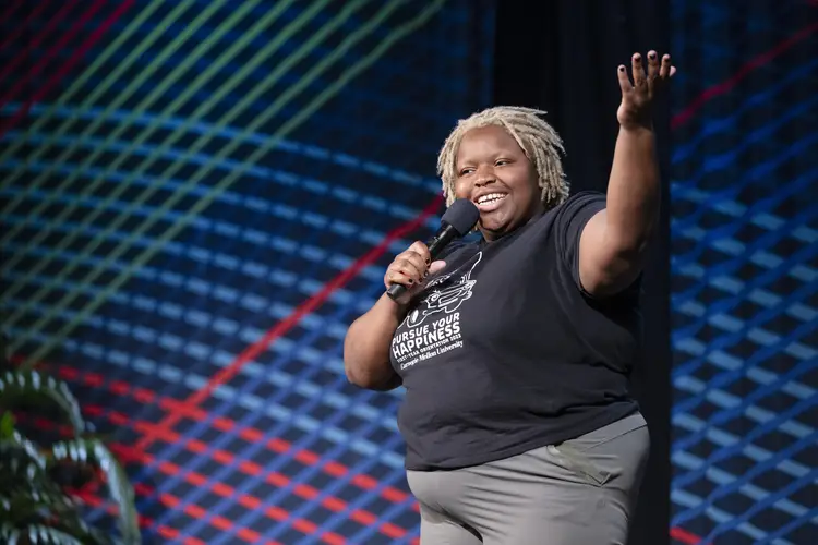 A Black woman with short blonde locks holds her arm up while she holds a microphone in her other hand and looks out toward the audience.