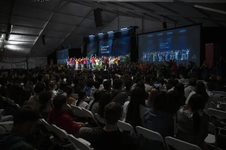 From a darkened audience, a crowd on a stage some distance away features people in bright solid-colored T-shirts gesturing while dancing together.