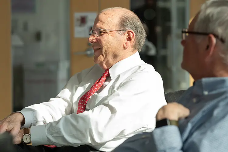 A man in a white dress shirt and a red tie is seen in profile with bright sun shining on him, as another man looks toward him from the right side of the photo.