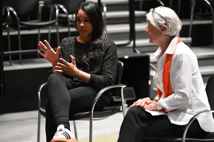 Two women seated in chairs look toward the left and speak to a crowd that is out of frame. The woman on the left is gesturing with her hands.