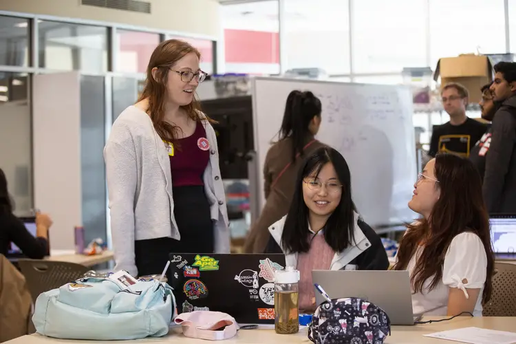Woman standing at edge of table talks to two female students sitting at the table in front of their laptops.