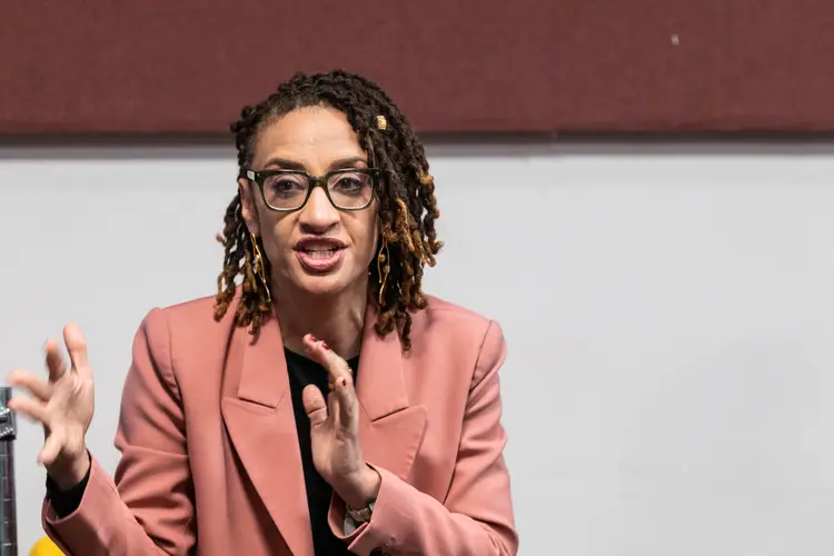 A woman with glasses wearing a salmon-colored coat gestures and talks