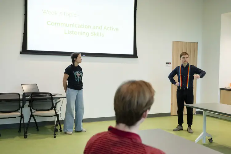 Two people stand at the front of a classroom underneath a projector screen talking while a student, listening and looking toward them, is framed in the foreground.
