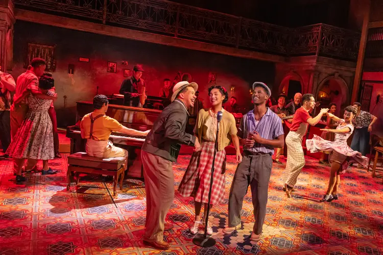 Two men and a woman stand around a microphone in the front of the photo, while behind them, in red-tinted light, other people dance and play instruments.