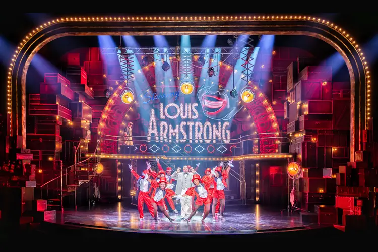 Stage and proscenium of a theater with sign on back wall that says "Louis Armstrong" and beams of light shining down on to cast members in red and silver sequined formal outfits in a small group.