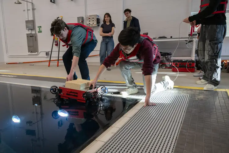 Two students in red live vests crouch near the corner of what looks like a swimming pool deck, each holding on to a black handle on a square red robot, as they drop it in to the water with their teammates behind them looking on.